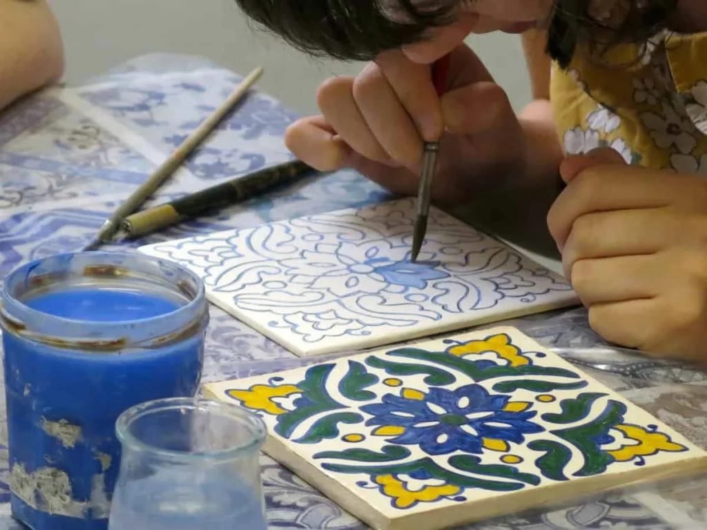 Person painting a traditional Portuguese azulejo tile by hand, surrounded by colourful ceramic paints and brushes on a workshop table